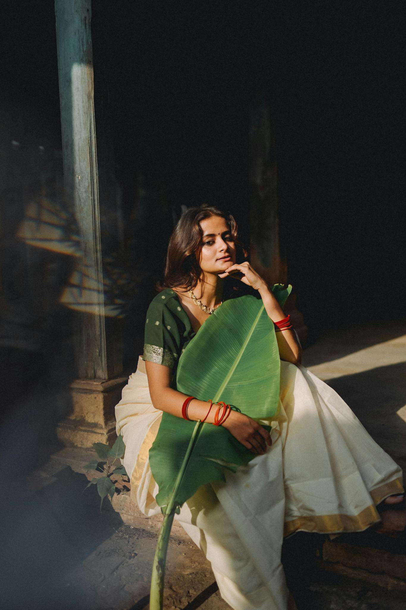 A woman in traditional Indian attire sits gracefully holding a large green leaf, embodying serene elegance.